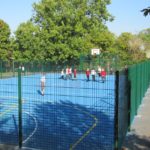 A group of children in sports kit play on an outdoor blue basketball court, part of a vibrant Multi-Use Games Area surrounded by green fencing and trees.