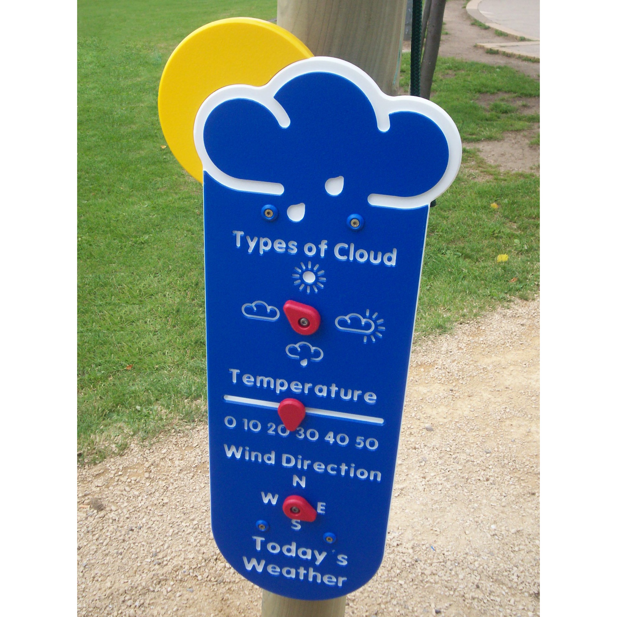 Blue playground weather panel with movable dials for cloud types, temperature, and wind direction. The panel is mounted outdoors on a post with grass and gravel path nearby.