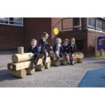 Five young children in school uniforms sit on wooden log play structures outdoors near a brick building, appearing to play and interact with one another.