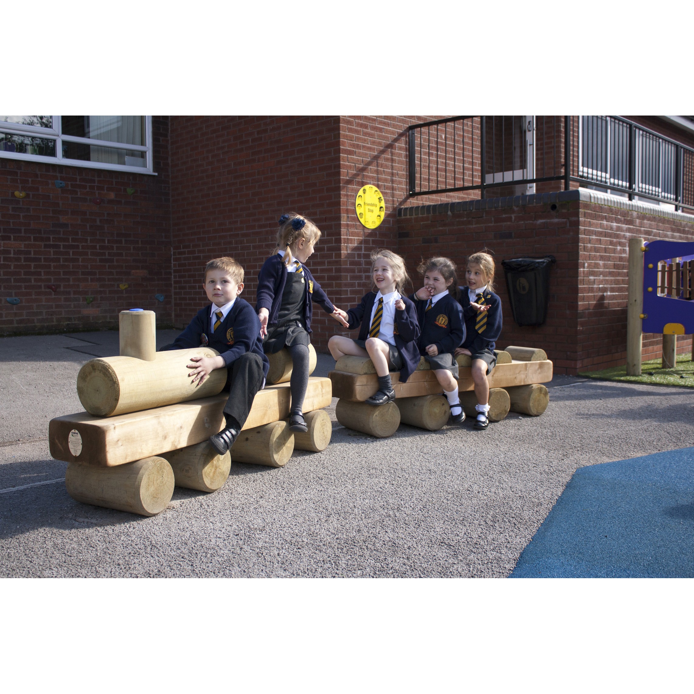 Five young children in school uniforms sit on wooden log play structures outdoors near a brick building, appearing to play and interact with one another.
