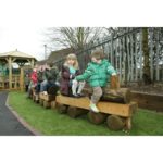 A group of young children sit on a wooden playground structure shaped like a train, outdoors near a fence and trees.