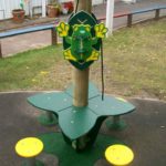 A green and yellow frog-shaped interactive panel is mounted on a wooden post above a green and yellow playground structure with seats, set outdoors.