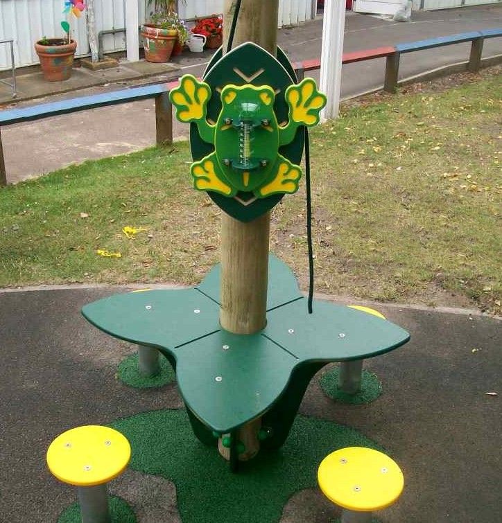 A green and yellow frog-shaped interactive panel is mounted on a wooden post above a green and yellow playground structure with seats, set outdoors.
