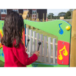 A child in a red shirt plays an outdoor musical instrument with metal tubes and colourful decorations at a playground.