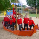 A group of schoolchildren in red jumpers stand and pose on a wooden playground structure shaped like a Viking ship.