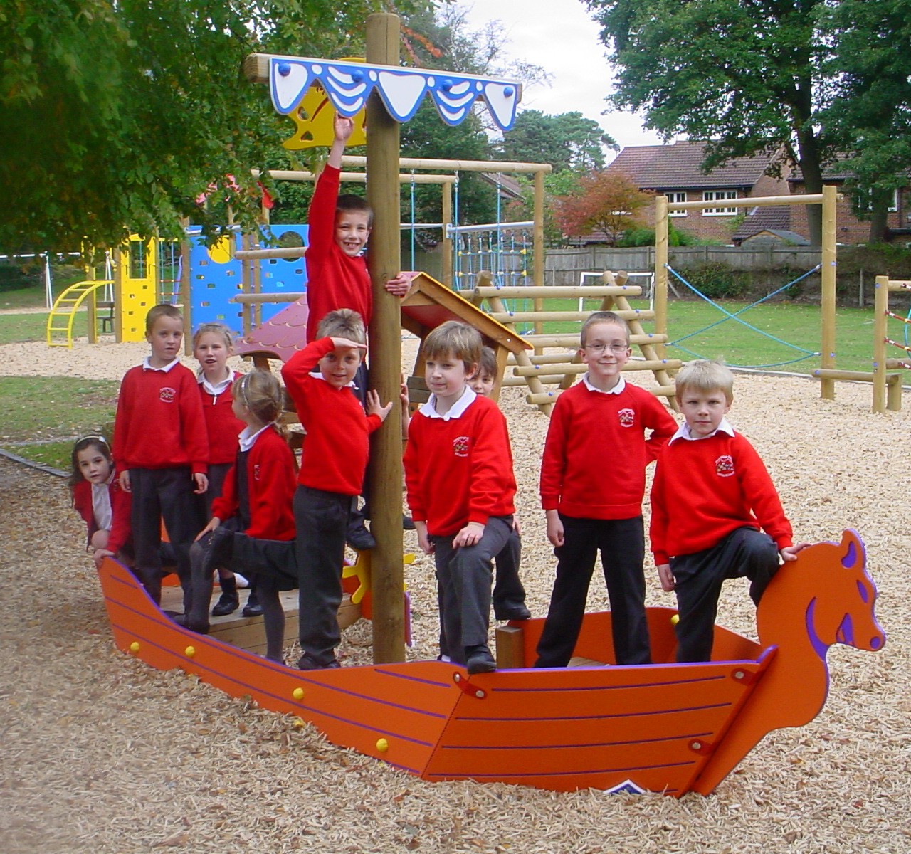 A group of schoolchildren in red jumpers stand and pose on a wooden playground structure shaped like a Viking ship.