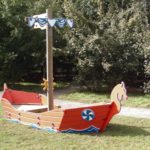Wooden playground structure shaped like a Viking ship with painted details, set on grass near a path and trees in the background.