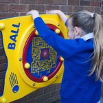 A child in a blue jumper interacts with a yellow and red wall-mounted ball maze game labelled BALL against a brick wall.
