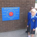 Three children in blue school uniforms stand near a wall-mounted learning clock with labelled times, used for teaching how to tell the time.