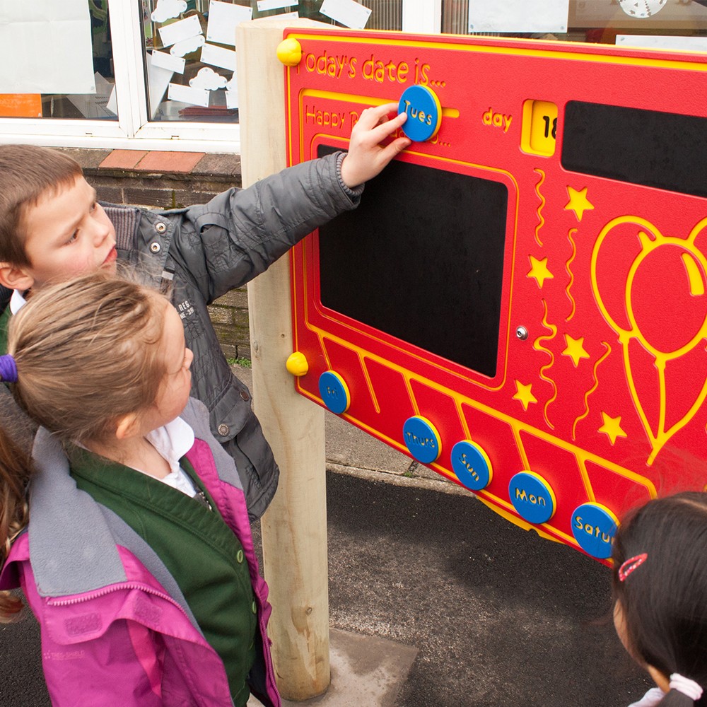 Three children interact with an outdoor educational board, with one child placing a circular label marked Tues on the day section.