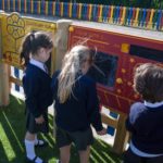 Three young children in school uniforms interact with educational boards outdoors on a playground with a colourful fence in the background.