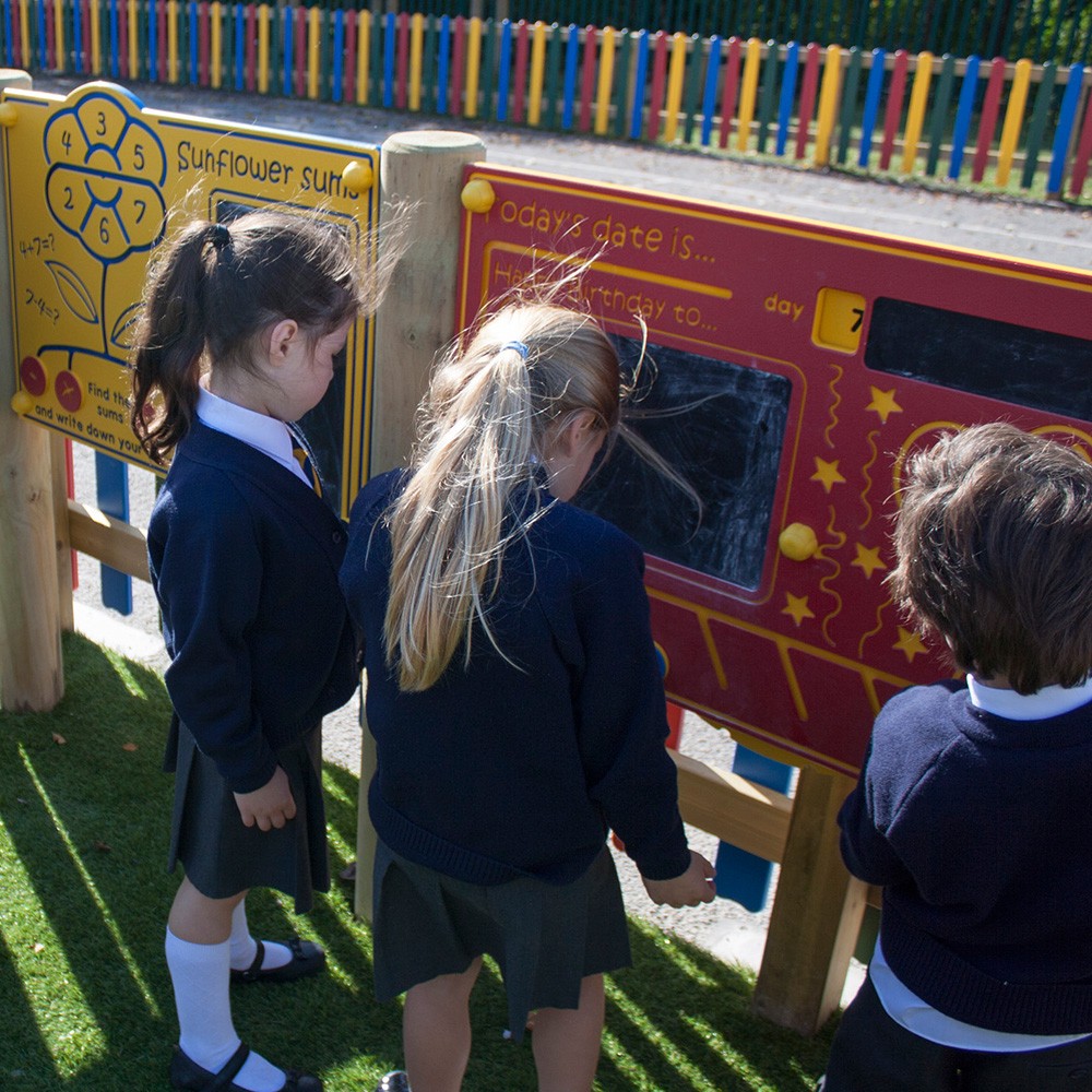 Three young children in school uniforms interact with educational boards outdoors on a playground with a colourful fence in the background.
