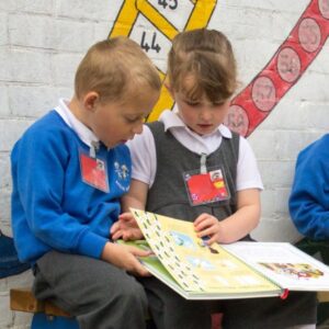 Two young children in school uniforms sit together and read a picture book, both wearing name badges, with a painted wall behind them.