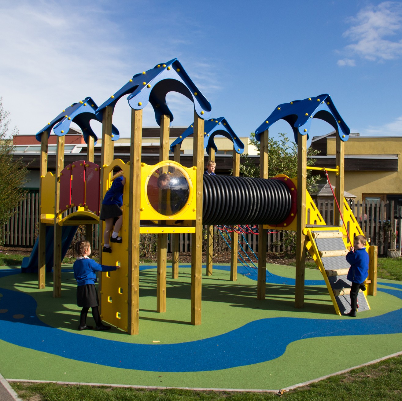 Children in blue uniforms play on a colourful playground structure with slides, climbing walls, and a tunnel, set on a blue and green rubber surface under a clear sky.