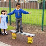 A boy balances on a springy playground board whilst two other children stand nearby, all dressed in school uniforms, with a fence and grass in the background.