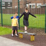 Three children in school uniforms stand by outdoor play equipment; one is balancing on a spring-mounted bench whilst the other two watch.