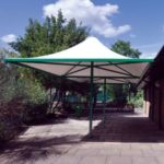 A white canopy with a green frame provides shade over a paved outdoor area near trees and a brick building under a partly cloudy sky.