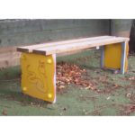 A wooden bench with yellow side panels, one featuring a cartoon snake illustration, stands on green outdoor flooring with scattered leaves.