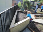Several young children stand beside a 75cm Log Planter filled with soil, some digging, in an outdoor area with adults nearby.