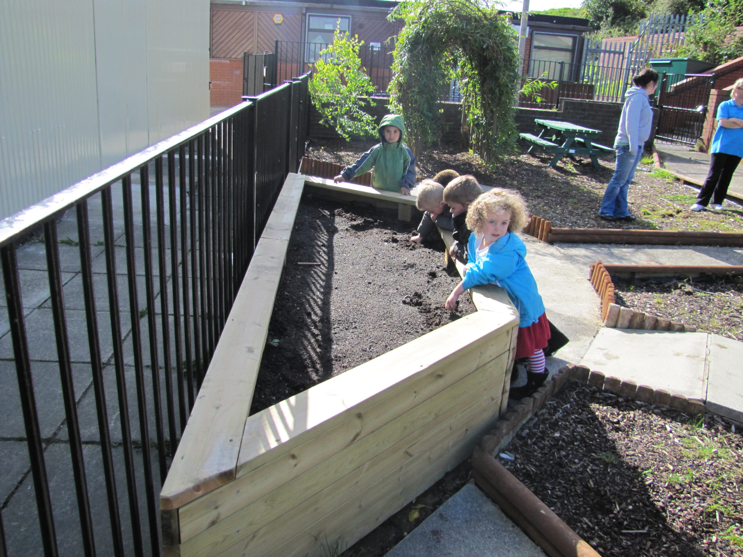 Several young children stand beside a 75cm Log Planter filled with soil, some digging, in an outdoor area with adults nearby.