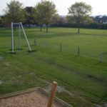 A grassy park with scattered trees, a metal swing frame without seats, wooden posts, and a mulched play area in the foreground.