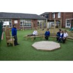 A teacher sits on a wooden chair whilst five children in school uniforms sit on curved benches in an outdoor classroom area.