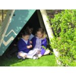 Three young children sit together on the grass inside a small outdoor tent, surrounded by greenery and sunlight.