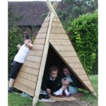 Three children play with a wooden outdoor tipi structure; two sit inside while one stands outside, smiling and posing. Lush greenery and a house are in the background.