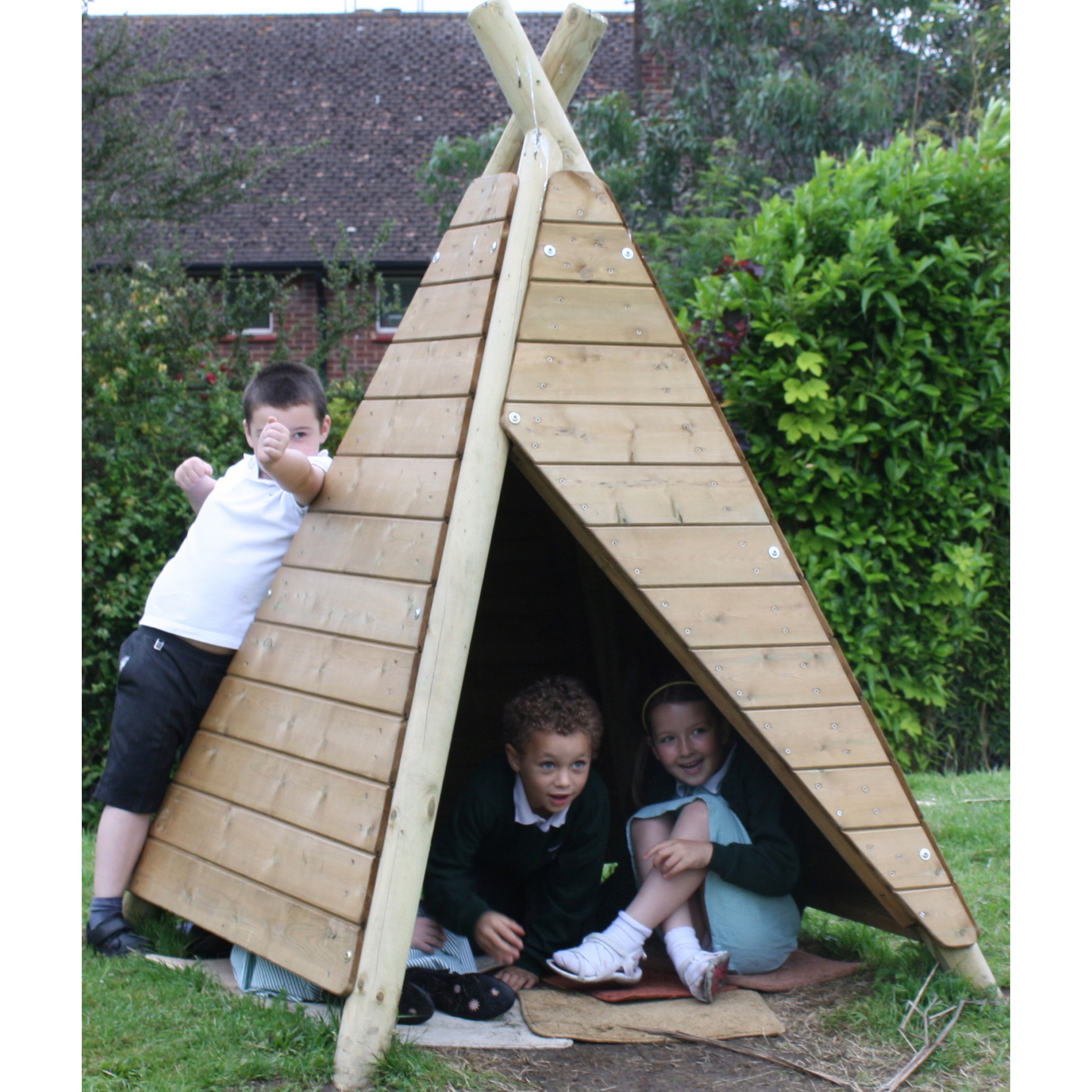 Three children play with a wooden outdoor tipi structure; two sit inside while one stands outside, smiling and posing. Lush greenery and a house are in the background.