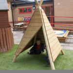 Two children sit inside a wooden tipi-style play structure on artificial grass in a school playground area.