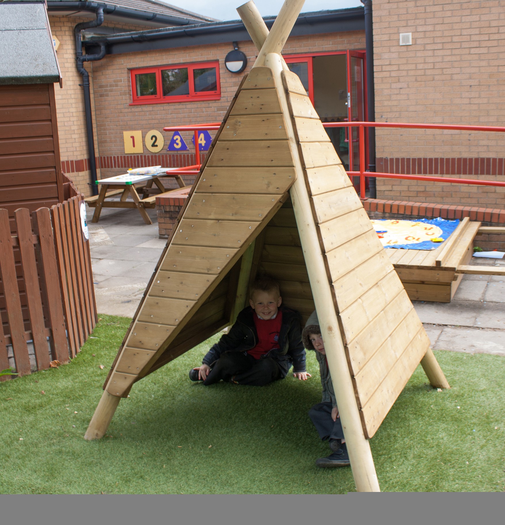 Two children sit inside a wooden tipi-style play structure on artificial grass in a school playground area.