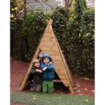 Three children in jackets crouch and smile inside a small wooden triangular hut outdoors, surrounded by greenery and a fence.