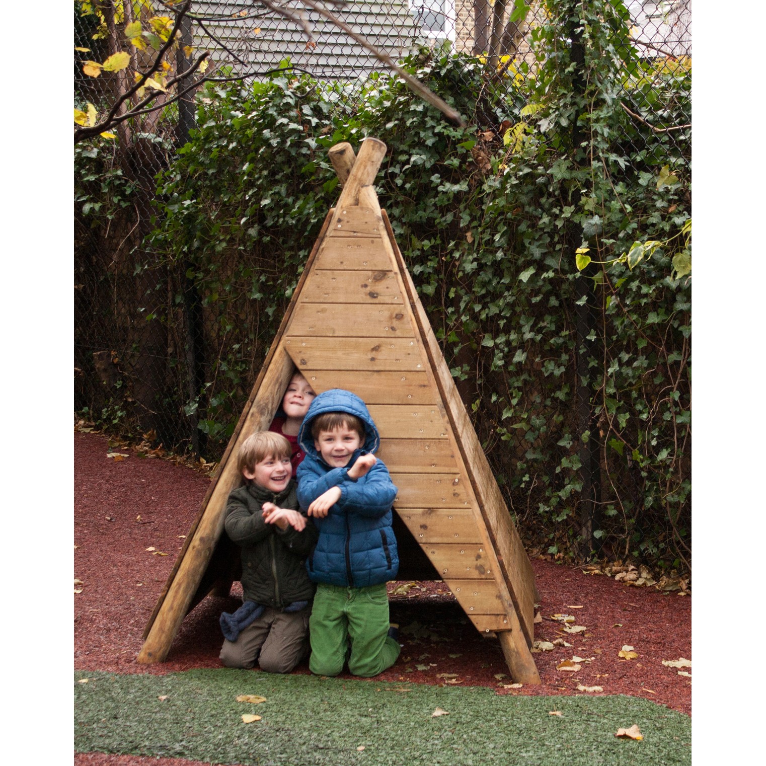 Three children in jackets crouch and smile inside a small wooden triangular hut outdoors, surrounded by greenery and a fence.