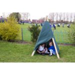 Three children sit inside a small makeshift tent on grass, whilst other children play in the background on a school field.