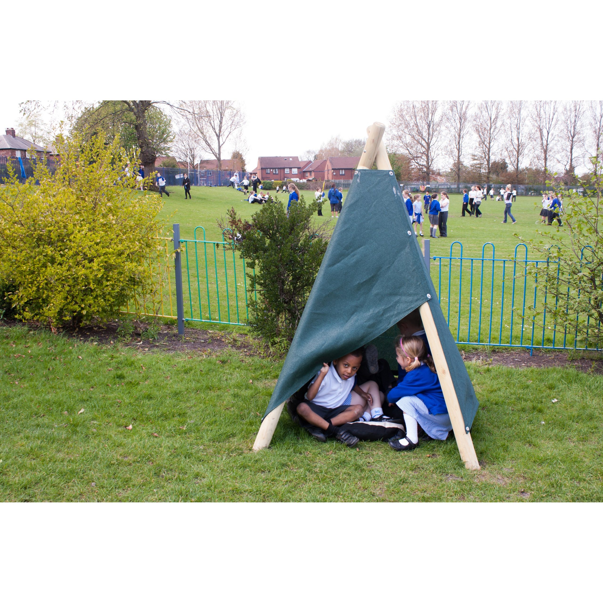 Three children sit inside a small makeshift tent on grass, whilst other children play in the background on a school field.