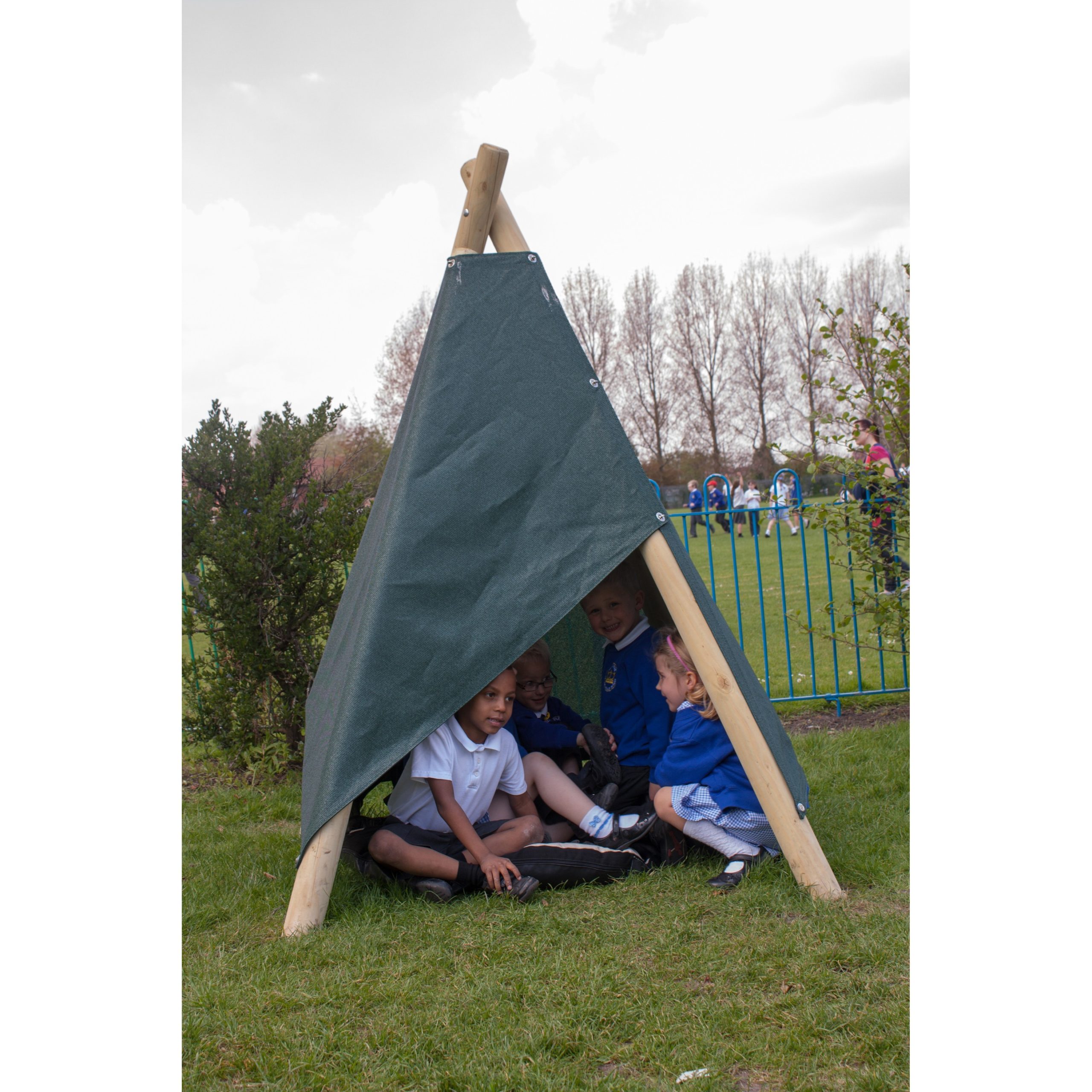 Four children sit under a small outdoor wooden and fabric shelter on grass, with bushes, trees, and a playground visible in the background.