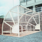 A white metal and glass bike shelter with racks for bicycles is positioned on a paved area near a building with a stone retaining wall.