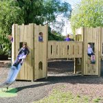 Five children play on the Fort Cumberland with Steel Slide, a wooden playground structure with towers, a bridge, climbing features, and a slide, surrounded by trees and wood chip ground cover.