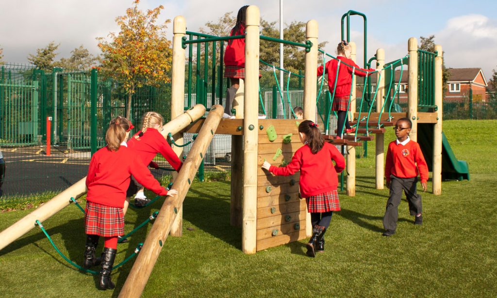 Children in red school uniforms play on a wooden climbing frame at a playground with artificial grass and fencing, discovering how to use (and get the most from) sensory play equipment as they explore and have fun.