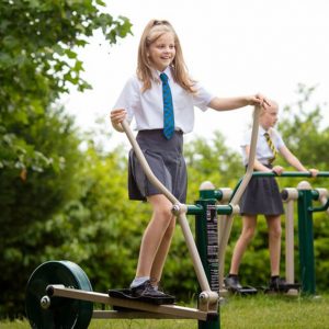 Two schoolgirls in uniform use outdoor exercise equipment in a green park area, one on a cross-trainer and another on a step machine.