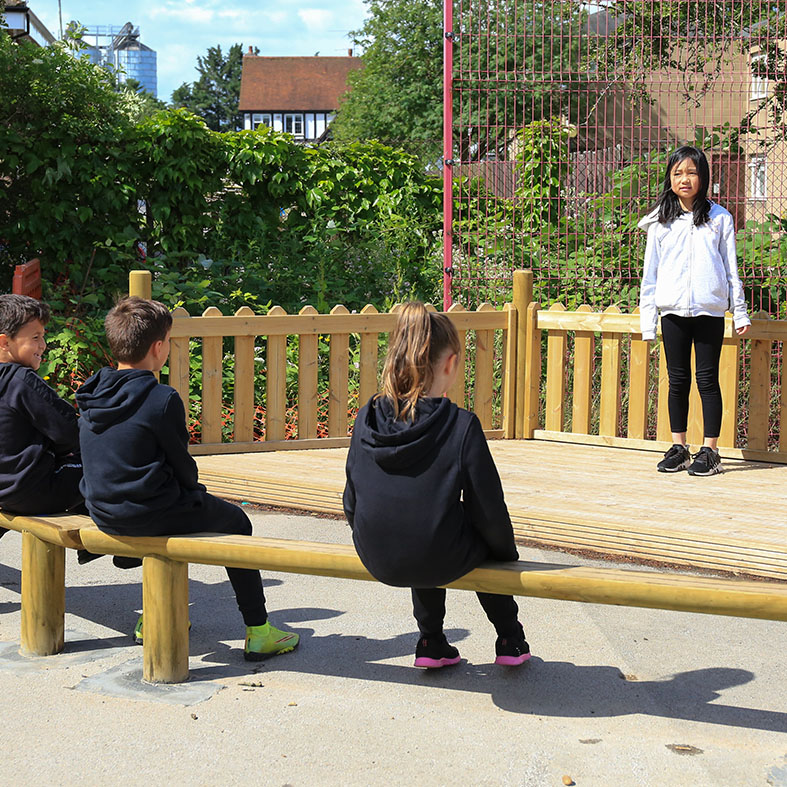 Four children are outdoors; three sit on a wooden beam while one stands on a platform in front of them, possibly addressing or performing for the group.