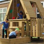 Three children play in a wooden playground structure with a sandpit in front of a brick building.