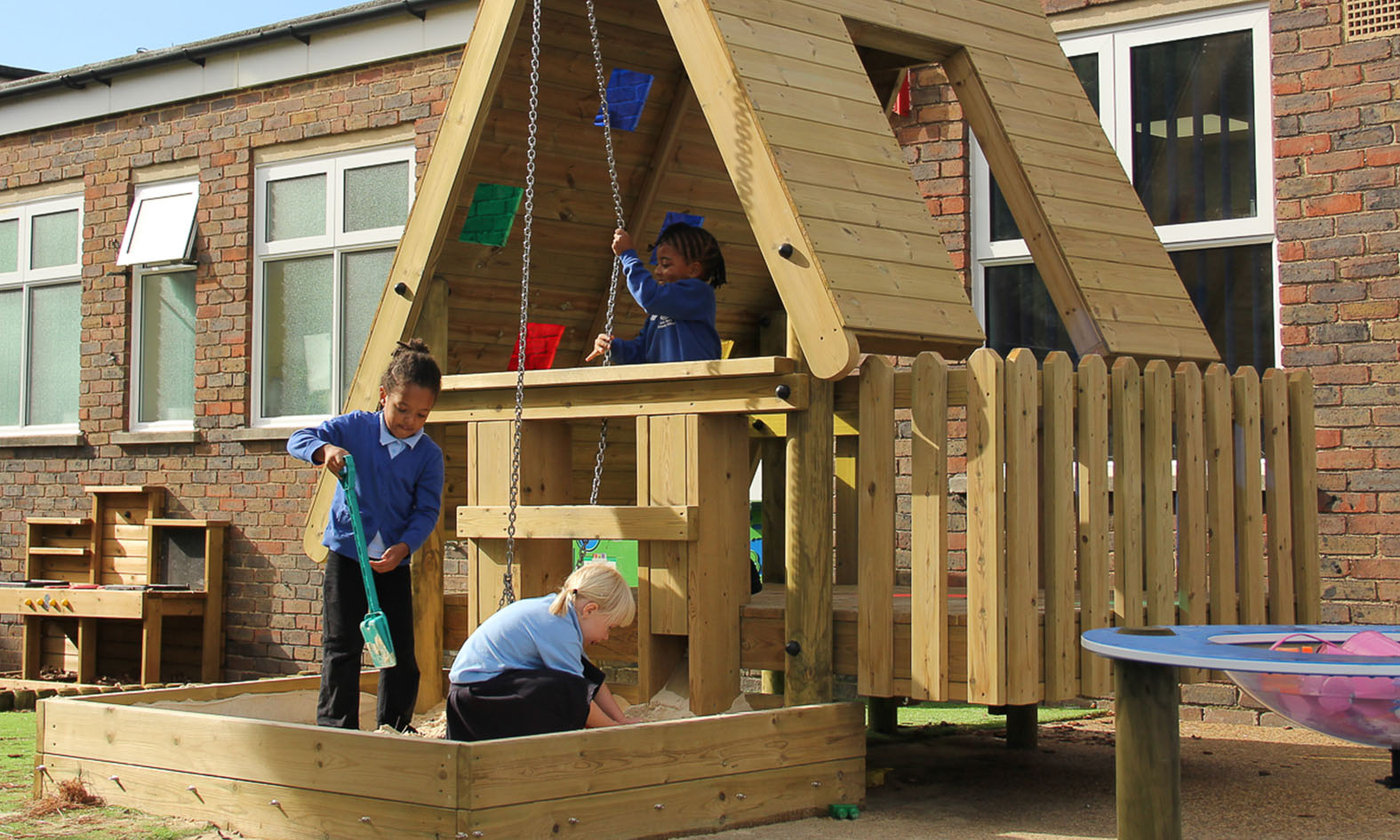 Three children play in a wooden playground structure with a sandpit in front of a brick building.