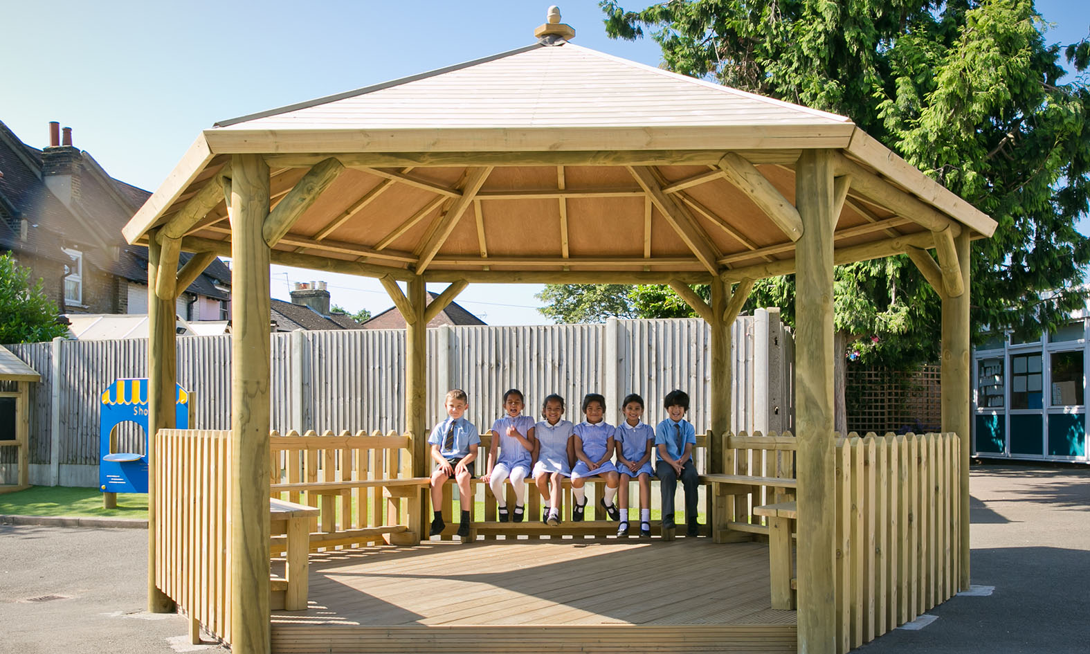 Six children in school uniforms sit on a bench inside a wooden gazebo—an example of all-weather play solutions for schools—on a playground, with a fence and trees in the background.