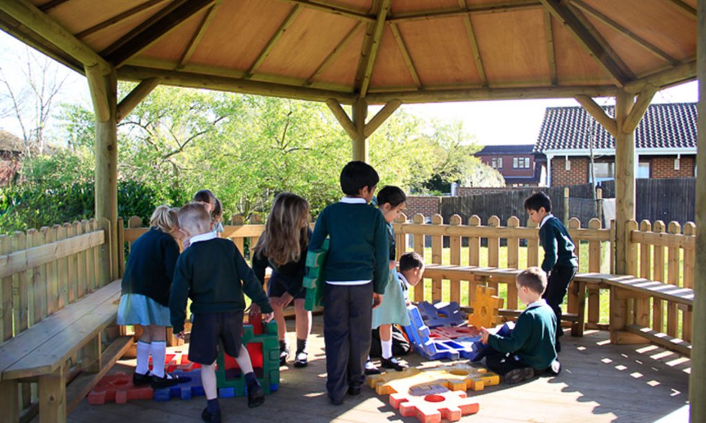 A group of young children in school uniforms assemble large foam puzzle pieces under a wooden gazebo in a fenced outdoor area.