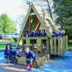 A group of children in jackets sit and stand on a wooden playhouse and sandpit in an outdoor playground surrounded by trees.