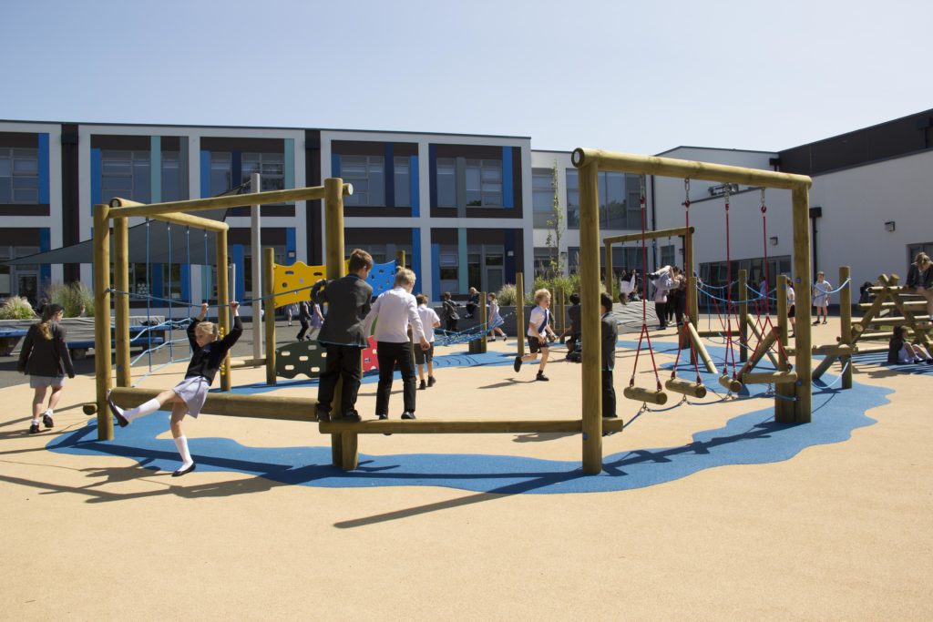 Children play on a wooden climbing structure in a school playground, maximising outdoor spaces, with a modern building in the background under a clear sky.