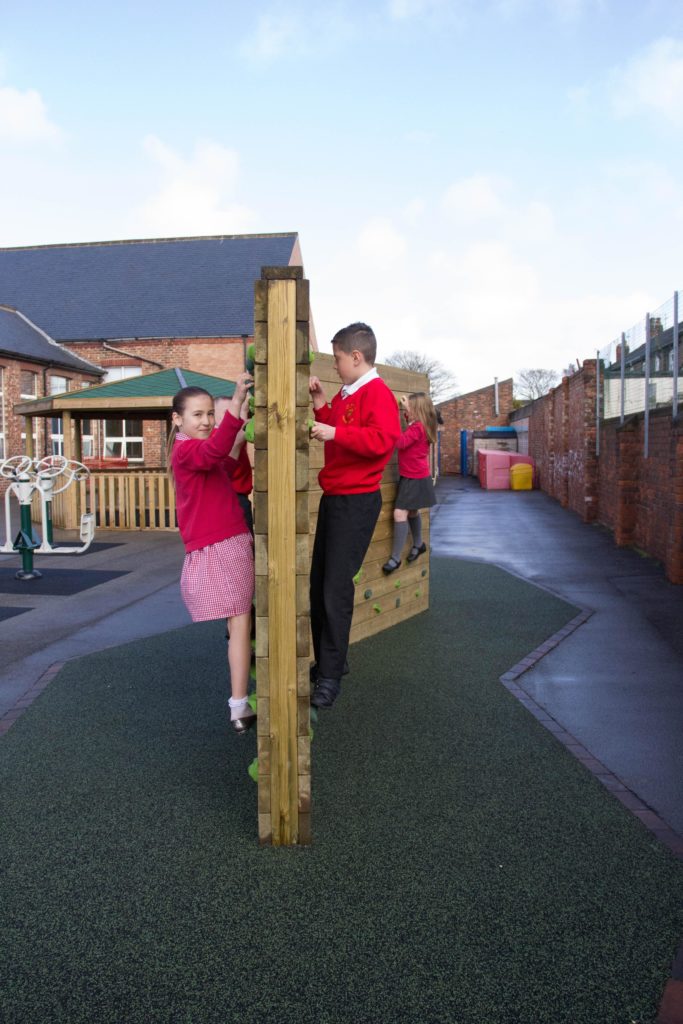 Two children in red school uniforms climb opposite sides of a well-maintained wooden climbing wall, highlighting the importance of maintaining playground equipment, with school buildings visible in the background.