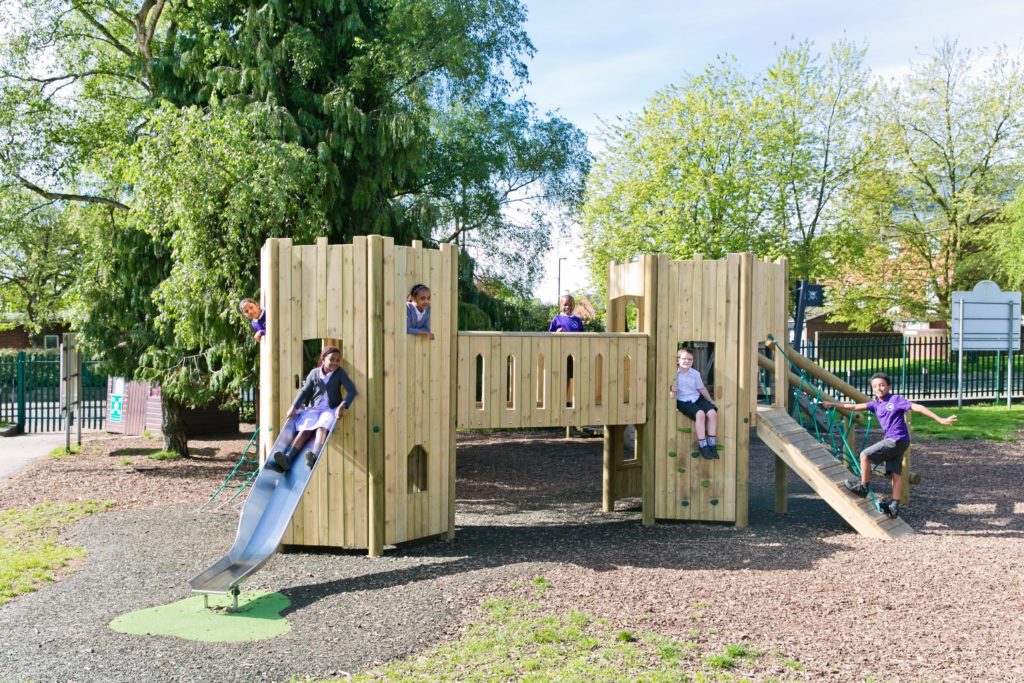Children play on Our Fort Range wooden playground structure with a slide, climbing wall, and bridge, surrounded by trees and a fence on a sunny day.