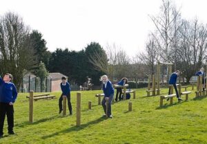 Several children in blue uniforms play on wooden balance beams and posts in a grassy outdoor playground area, enjoying activities that support children's mental and physical health on a sunny day.
